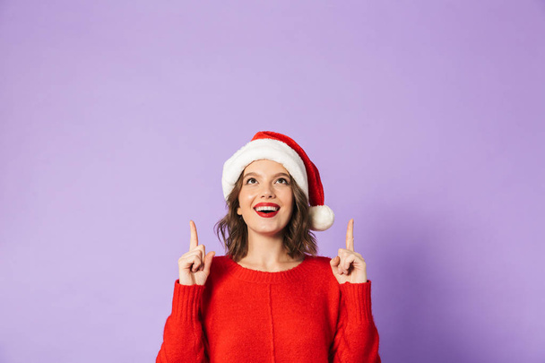 Portrait of an excited happy young woman wearing christmas hat isolated over purple background pointing. - Foto, Imagem