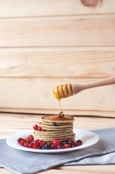 Een stapel pannenkoeken met bessen en honing op een witte plaat en keuken attributen. - Foto, afbeelding