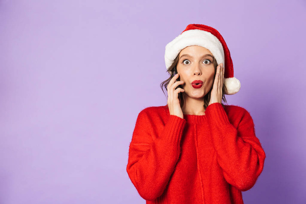 Portrait of an excited happy young woman wearing christmas hat isolated over purple background talking by mobile phone. - Фото, зображення