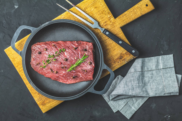 Fresh raw meat beef steak with ingredients for cooking spices, thyme and rosemary in black frying pan on dark background, top view, copy space for you text. - Foto, immagini