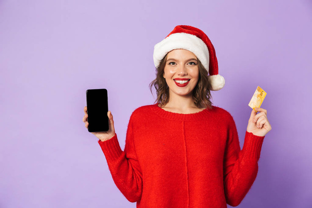 Portréja egy boldog fiatal nő, fárasztó karácsonyi kalap, elszigetelt, felett lila háttér gazdaság hitelkártya mobiltelefon használata. - Fotó, kép