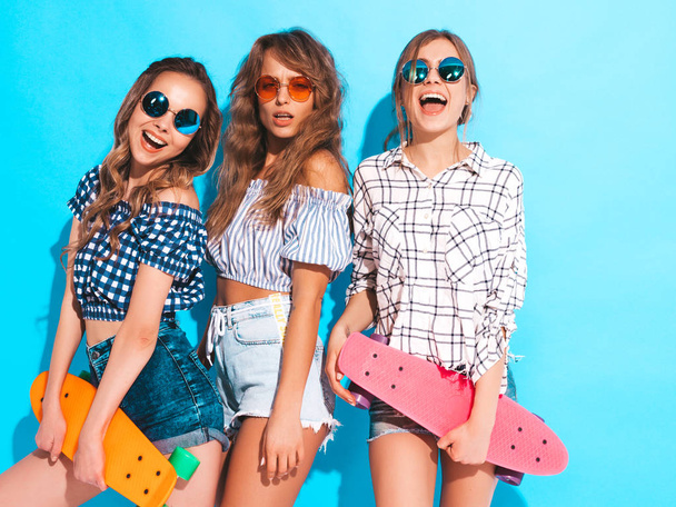Three young stylish sexy smiling beautiful girls with colorful penny skateboards. Women in summer hipster checkered shirt clothes isolated on blue wall in sunglasses. Positive models having fun  - Foto, Bild
