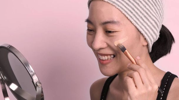 Chica asiática o mujer de 40 años de edad hermosa cara con aspecto japonés que componen por líquido de base y cepillo cosmético en la piel sensible para ayudar a su tez mirada impecable y sin retoque. - Foto, imagen