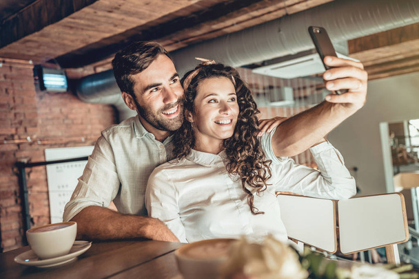 Juntos! Hermosa pareja haciendo selfie en el nuevo teléfono inteligente. Pareja enamorada pasa tiempo en la cafetería
. - Foto, Imagen