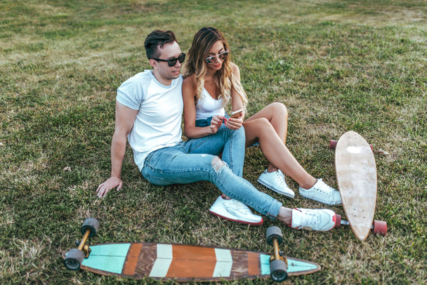 A young and beautiful couple man a woman, in summer park, stand grass, in hands of smartphone, chatting on Internet, near skateboard and longboard for skating. In casual clothes and sunglasses. - Foto, imagen