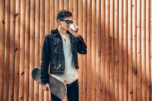 Young stylish man with skateboard and drinking coffee standing near wooden background and looking away.Lifestyle concept. - Foto, afbeelding