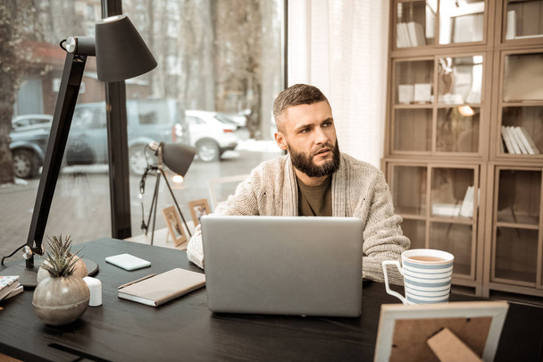 Bearded man sitting in grey cardigan with giant windows on the background - Foto, imagen