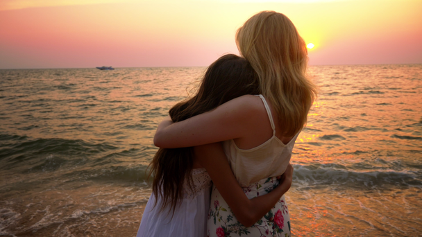 μητέρα και κόρη στην άσπρα φορέματα περπατάτε ξυπόλητοι σε μια αμμώδη παραλία, κρατώντας τα χέρια με φόντο ένα μαγευτικό ηλιοβασίλεμα - Πλάνα, βίντεο