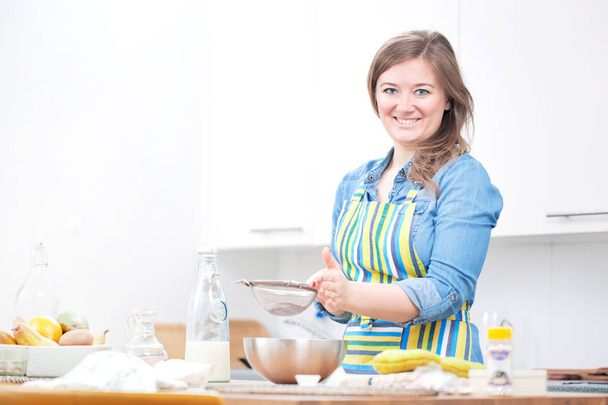cheerful woman in apron sifting flour and smiling at camera in kitchen - Foto, Imagem