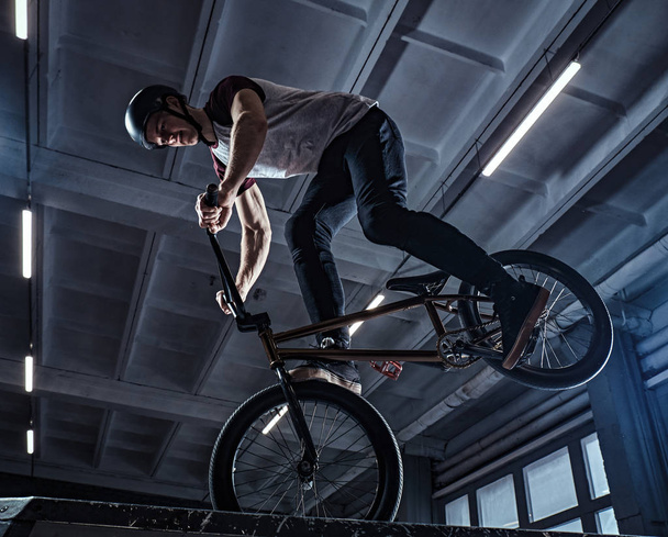 Молодой человек с BMX выполняет трюки в скейтпарке в помещении
 - Фото, изображение