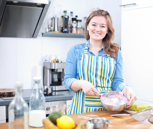 fröhliche junge Frau in Schürze, die Eier verquirlt und in der Küche in die Kamera lächelt  - Foto, Bild