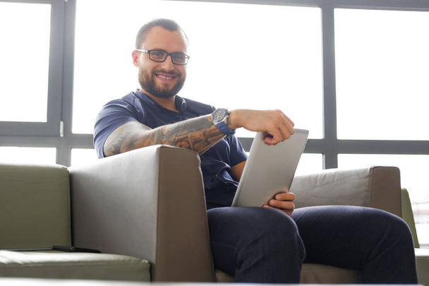 Homme d'affaires prospère vêtu d'une chemise bleue assis sur le canapé dans le bureau, tenant une tablette, souriant et regardant une caméra
 - Photo, image