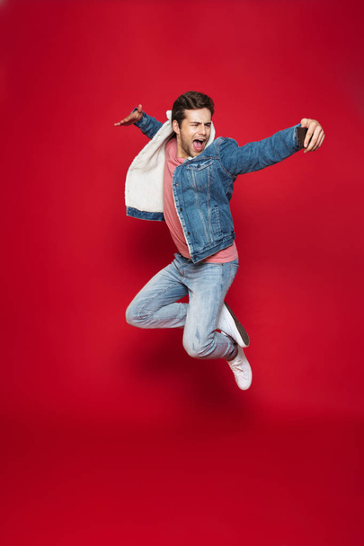 Full length of a cheerful young man wearing warm denim jacket jumping isolated over red background, taking a selfie - Foto, Imagen
