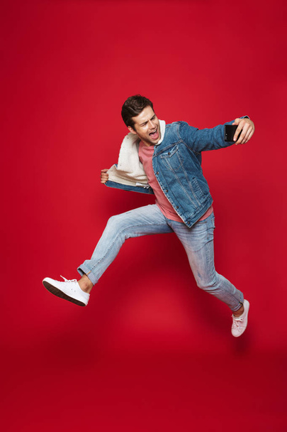 Full length of a cheerful young man wearing warm denim jacket jumping isolated over red background, taking a selfie - Valokuva, kuva