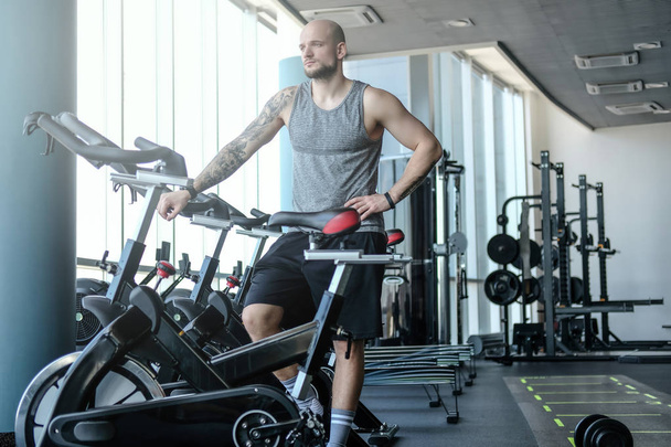 Retrato de un hombre atlético calvo con un tatuaje en la mano parado junto a una bicicleta estática en el moderno gimnasio
 - Foto, Imagen