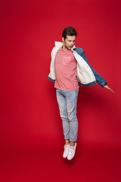 Full length of a happy young man wearing warm denim jacket jumping isolated over red background - Foto, imagen