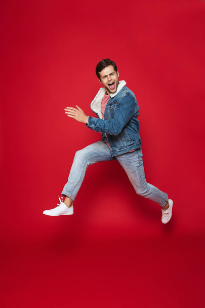 Full length of a happy young man wearing warm denim jacket jumping isolated over red background - Foto, immagini