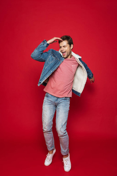 Full length of a happy young man wearing warm denim jacket jumping isolated over red background - Foto, Bild
