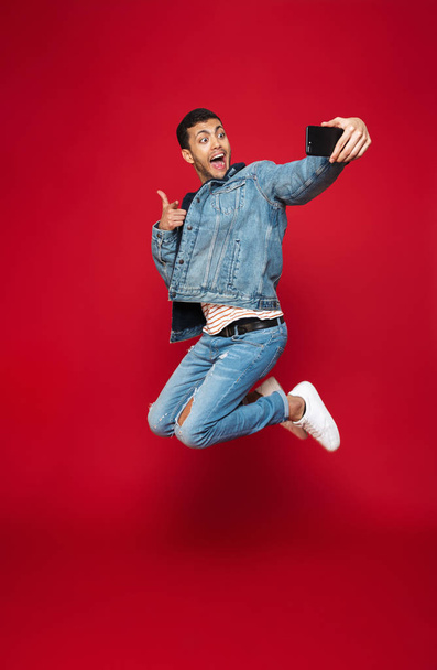 Full length of a cheerful young man wearing warm denim jacket jumping isolated over red background, taking a selfie - Φωτογραφία, εικόνα