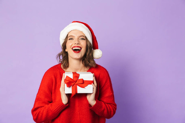 Cheerful young woman wearing red Christmas hat standing isolated over violet background, holding gift box - Fotó, kép