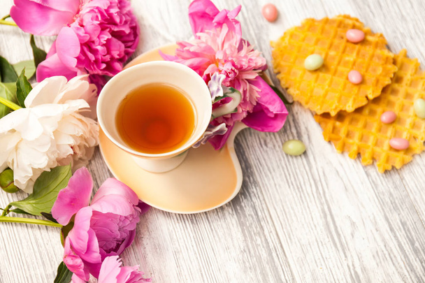 Filiżanka zielonej herbaty i wiosna blossom piwonie na stare drewniane tła z goframi i karmelki. Rustykalne. - Zdjęcie, obraz