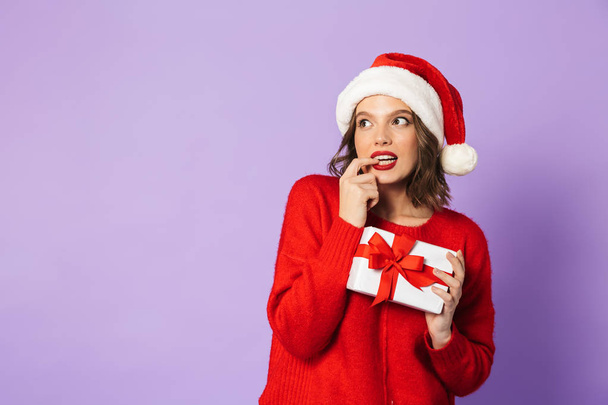 Smiling pensive young woman wearing red Christmas hat standing isolated over violet background, holding gift box - Foto, Imagem
