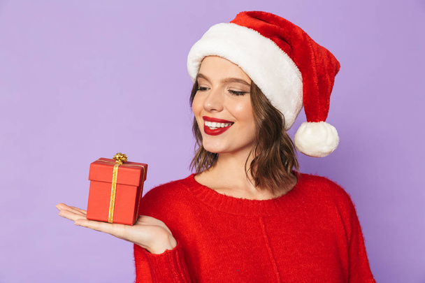 Portrait of an excited happy young woman wearing christmas hat isolated over purple background holding surprise gift box. - Fotó, kép