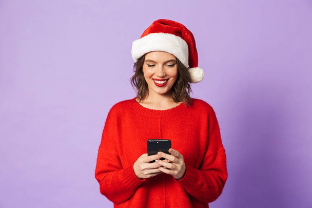 Portrait of a happy young woman wearing christmas hat isolated over purple background using mobile phone. - 写真・画像