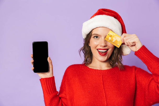 Portret szczęśliwy młodej kobiety sobie Boże Narodzenie kapelusz na białym tle nad fioletowym tle przy użyciu telefonu komórkowego z posiadania karty kredytowej. - Zdjęcie, obraz