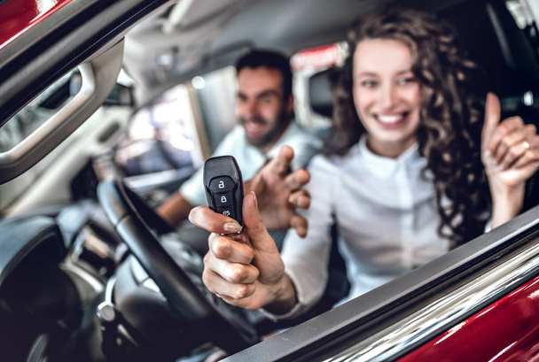 Close up of attractive woman showing car key through the window. Nice young couple sitting inside the new car at the dealership. Focus on key - Zdjęcie, obraz