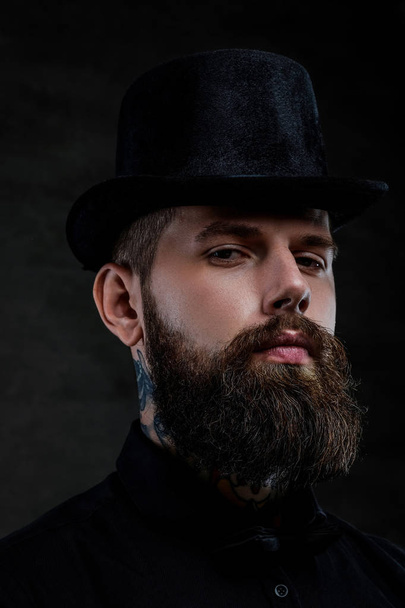 Крупним планом портрет старомодного бородатого чоловіка з татуюваннями на шиї в верхньому капелюсі, ізольованого на темному тлі. Студійний знімок
 - Фото, зображення