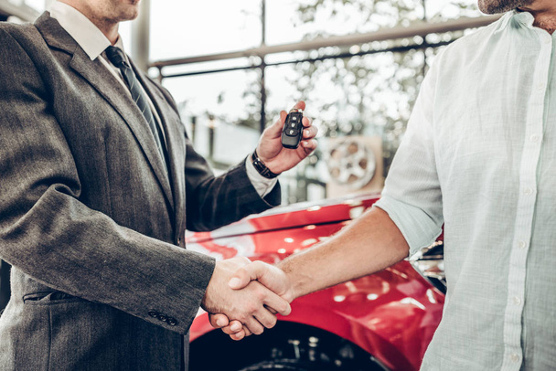 Auto business, car sale, deal, gesture and people concept - close up view of dealer giving key to new owner and shaking hands in auto show or salon - Foto, imagen