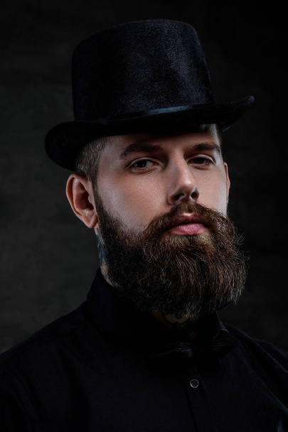 Retrato de close-up de um homem barbudo à moda antiga com tatuagens no pescoço usando um chapéu superior, isolado em um fundo escuro. Estúdio
 - Foto, Imagem