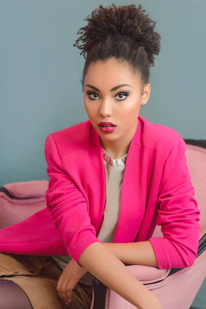 beautiful attractive young mixed rase woman with make up and in pink blazer sitting in the armchair - Photo, Image