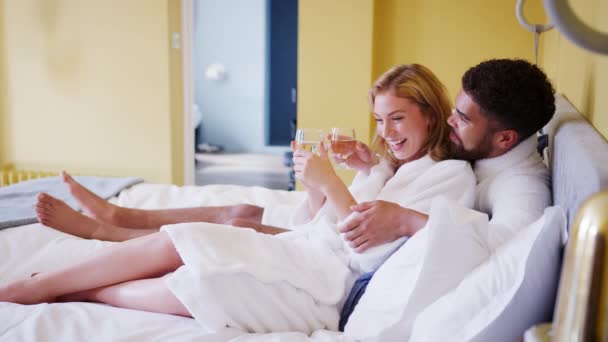Mixed race young adult couple reclining on the bed in a hotel room in bathrobes holding glasses of wine and talking, lockdown - Felvétel, videó