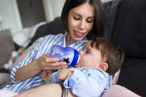 Shot of a pretty young mother feeding her baby with milk on feeding bottle in the living room of home. - Foto, afbeelding