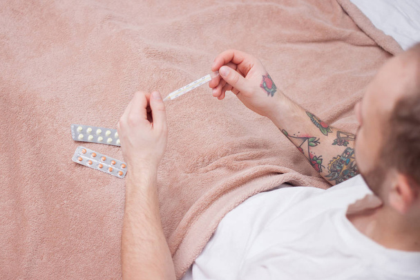 Sick man with tattoos on his arm lying on sofa checking his temperature  - Photo, image