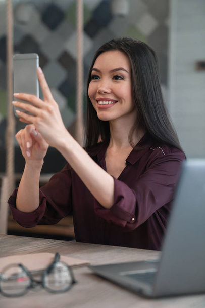 Attractive Asian woman in purple blouse smiling and making selfie on her phone while sitting at cafe - Photo, Image
