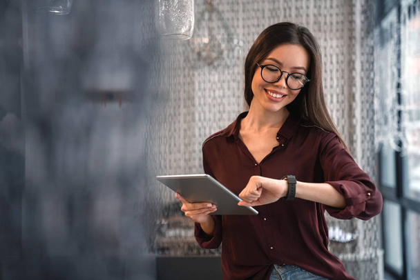 Portrait of business woman in eyeglasses checking time on watch and tablet on hand standing in the office - Foto, imagen