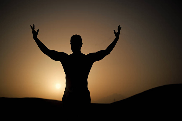Man with fit silhouette on sunset sky. Sportsman with raised hand on natural landscape. Athlete show power. Workout in summer dusk. Sport, wellness and bodycare concept - Photo, Image