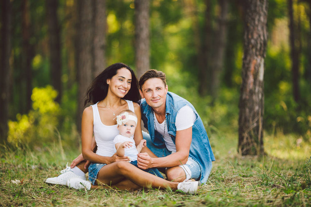 The theme family outdoor recreation in forest park area. Young beautiful stylish caucasian family with small child daughter one year old sit on the grass in the coniferous forest in summer at sunset. - Fotografie, Obrázek