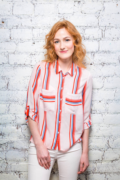 A beautiful young woman student with red curly hair and freckles is leaning against a brick wall of gray color. Dressed in a red striped shirt and white pants. - Foto, afbeelding