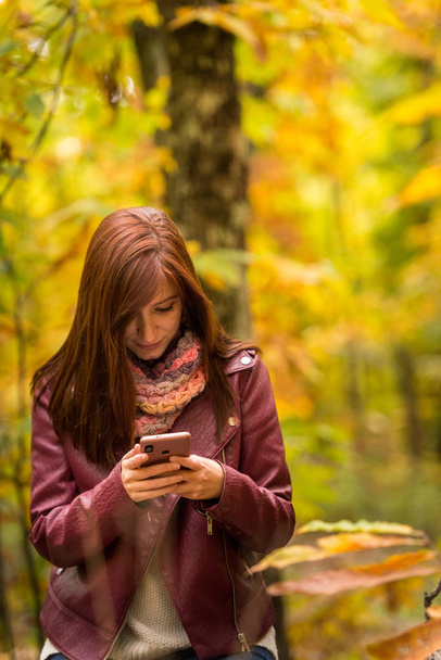 Молодая рыжая женщина смотрит на свой мобильный телефон в лесу
 - Фото, изображение