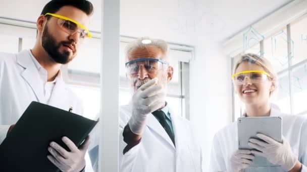 Group of chemists working in a lab. Young white male and female chemists with senior caucasian chemist working together in lab, writing on glass display. Science concept. - Кадри, відео