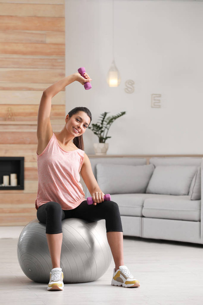 junge Frau turnt zu Hause mit Kurzhanteln auf Fitnessball - Foto, Bild