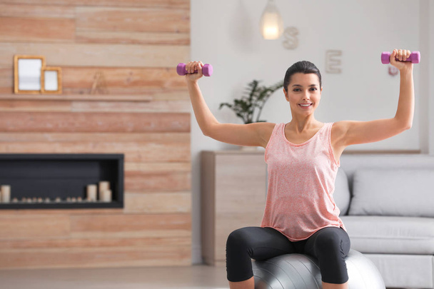 Young woman doing exercise with dumbbells on fitness ball at home. Space for text - Foto, Imagen
