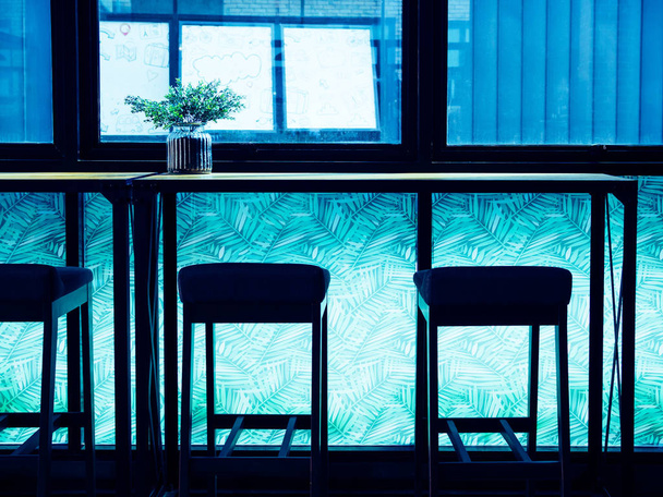 Sgabelli da bar Silhouette e fiore in vaso su bancone in legno bar vicino alla finestra vetro sulla stanza luce blu in caffè
. - Foto, immagini
