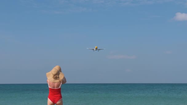 Toeristische vrouw in rode zwembroek en hoed verwelkomt vliegtuig voor reizen. Hallo vakantie concept - Video