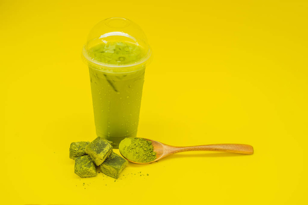Bir plastik kap ve saman ve tozu matcha sarı zemin üzerine kaşıkla buzlu yeşil çay latte. Ev yapımı Matcha Latte çayları ile süt sıfır atık - Fotoğraf, Görsel