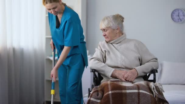 Rude hospital janitor cleaning room, moving wheelchair with old female patient - Video